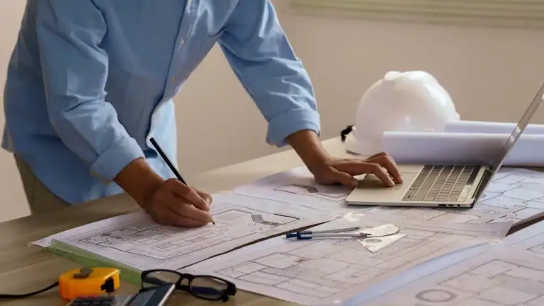 Detaljerte arkitekttegninger og designplaner på arbeidsbordet, med måleinstrumenter og en datamaskin, for å illustrere arkitekturprosessen og snittutforming.